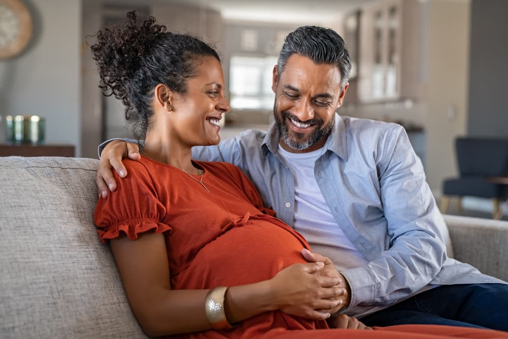 A couple sat together, the male partner is holding his pregnant female partner's pregnancy bump and smiling